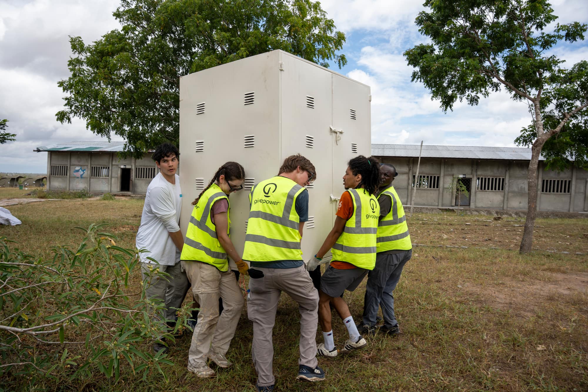 Trekkers in safety vests carrying a battery enclosure cabinet across a school field in Kenya